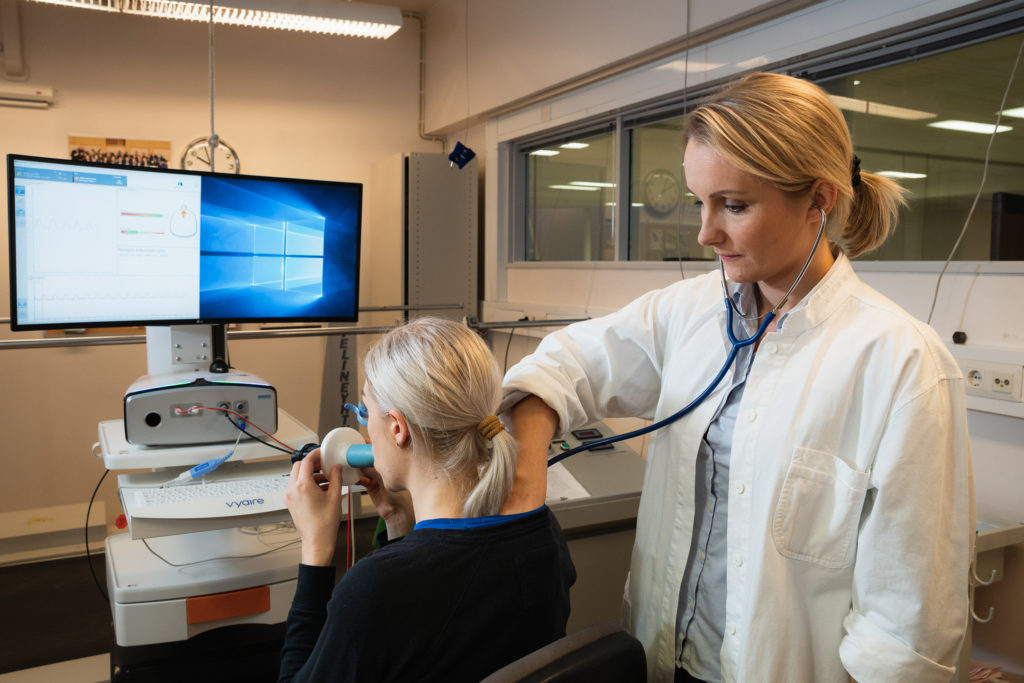 Liikuntalääketieteen erikoislääkäri Kerttu Toivo kuuntelee stetoskoopilla urheilijan keuhkoja. Urheilija istuu tuolilla ja tekee hengittää spirometriin.